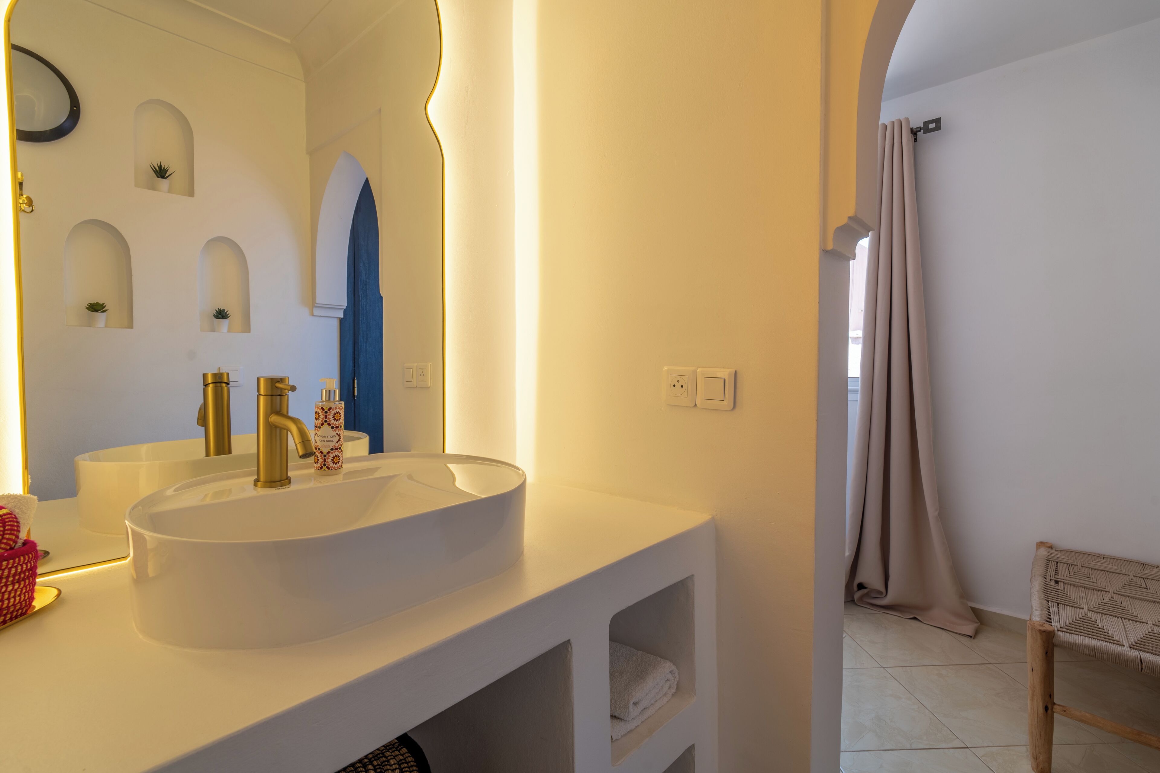 Un baño con lavamanos blanco, grifo dorado y espejo. Hay plantas en macetas en la pared y un perchero para toallas.
