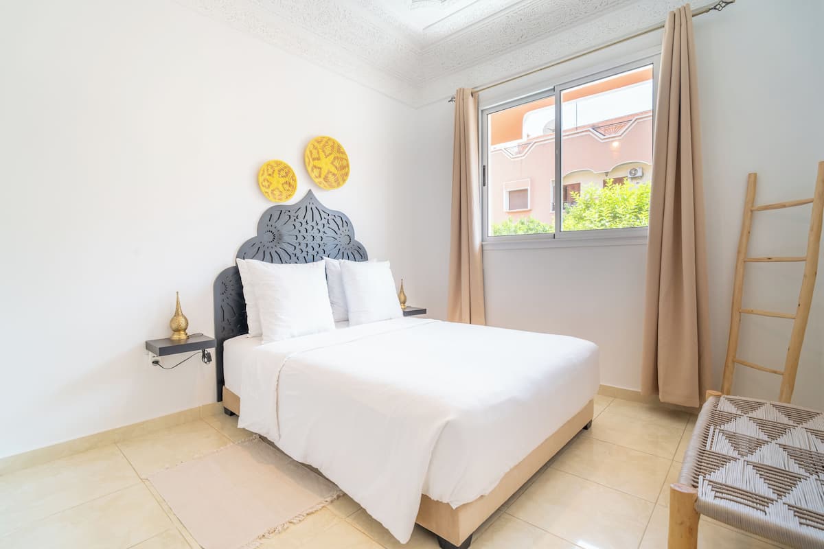 A bedroom with a bed, a nightstand, a window with curtains, and a view of buildings outside.