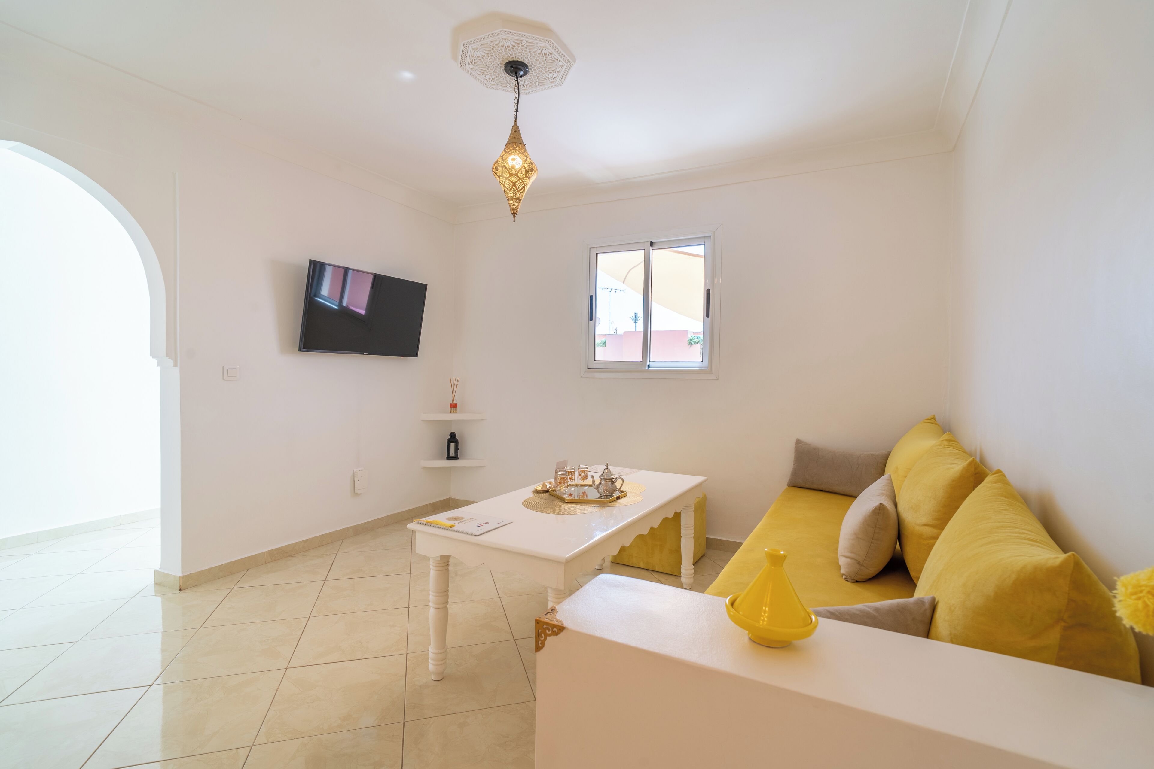 A modern living room with a white sofa, a coffee table, a TV mounted on the wall, and a window providing natural light.