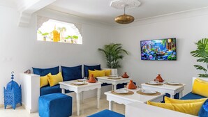 Een kamer met een kleurenschema in wit en blauw, met een zithoek, een eettafel met oranje aardewerk en een groot raam met uitzicht op palmbomen.
