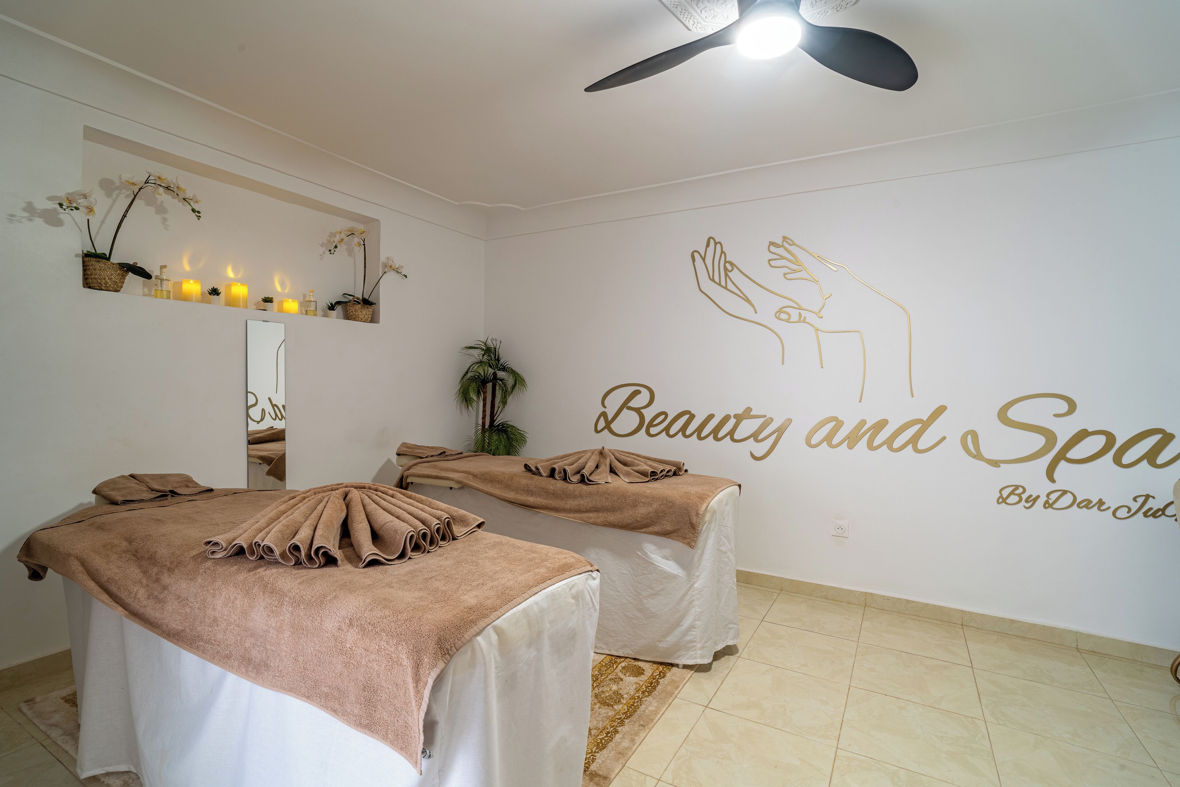 A spa room with two massage tables, a ceiling fan, a wall-mounted mirror, and a wall with the text "Beauty and Spa By Dar Tubs."