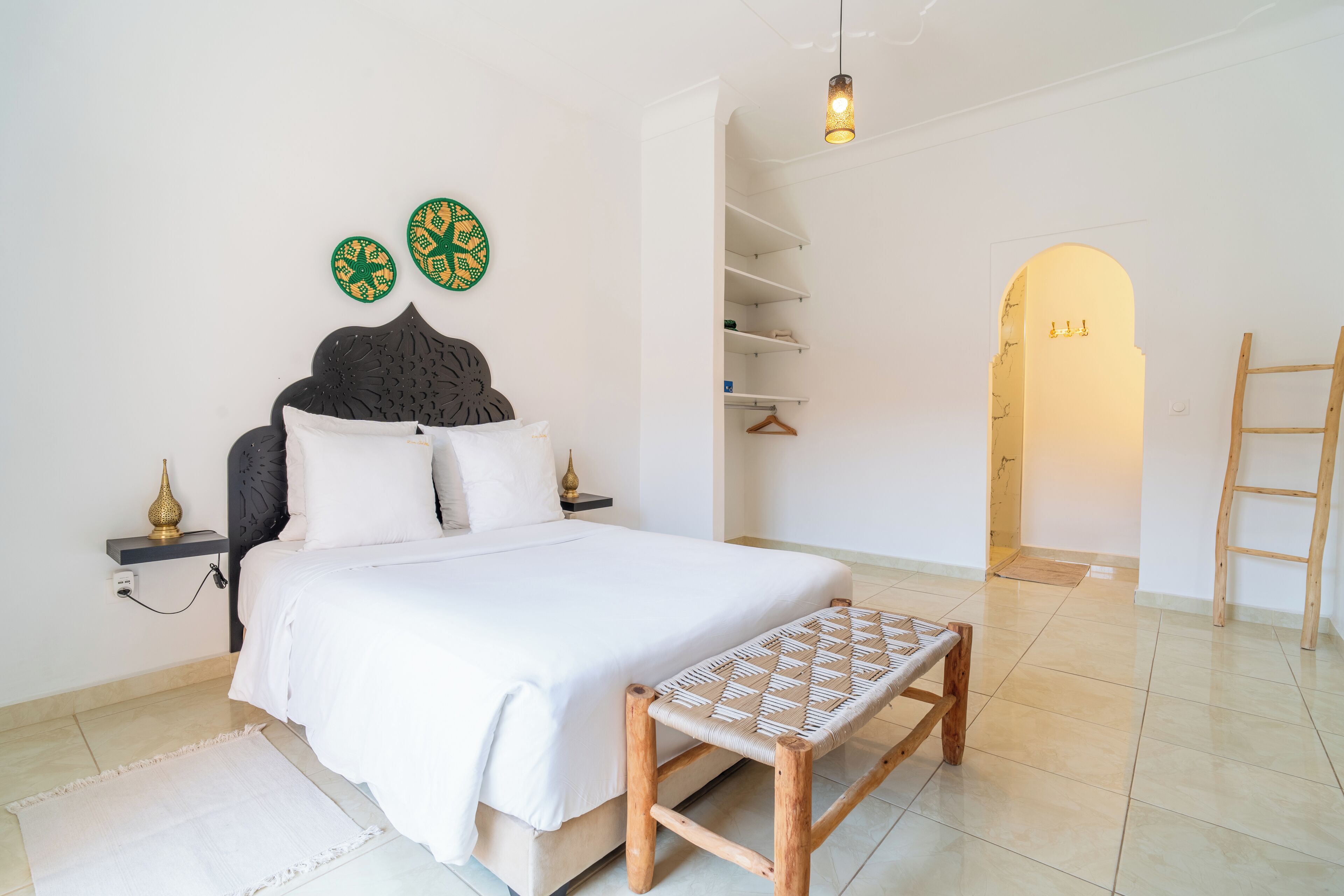 Une chambre à coucher comprenant un lit, des tables de chevet, un banc, une armoire et une porte donnant sur une autre pièce.
