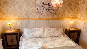 Un lit bien fait, avec une literie à motifs floraux, deux tables de chevet avec des lampes, une illustration de vélo accrochée au mur et une tête de lit en bois.