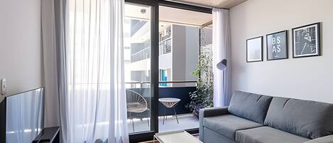 A modern living room with a grey sofa, a flat-screen TV, a small white table, and a sliding glass door leading to an outdoor balcony.