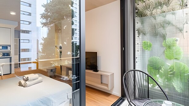 A modern bedroom with a bed, a TV, and a glass door leading to a balcony with a view of greenery. - Urban Rent - Departamentos en el corazon de Belgrano (Buenos Aires)