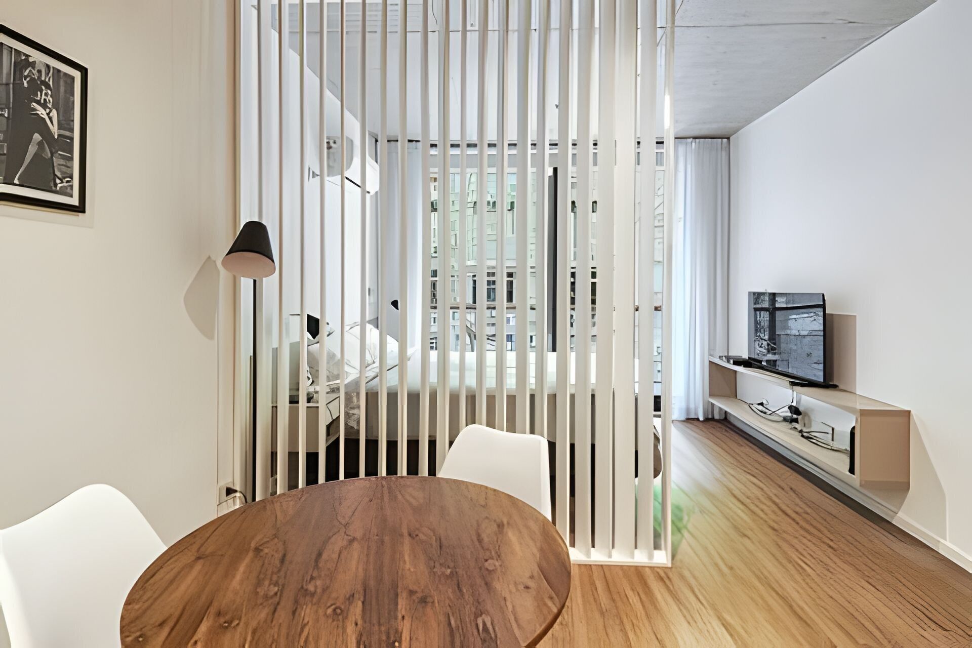 Un salon moderne avec une table à manger en bois, des chaises blanches, un téléviseur posé sur un meuble en bois et une cloison blanche agrémentée de stores verticaux.