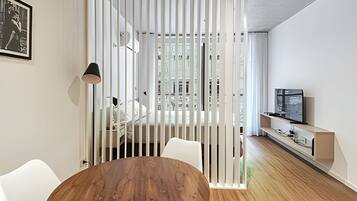 Un salon moderne avec une table à manger en bois, des chaises blanches, un téléviseur posé sur un meuble en bois et une cloison blanche agrémentée de stores verticaux.