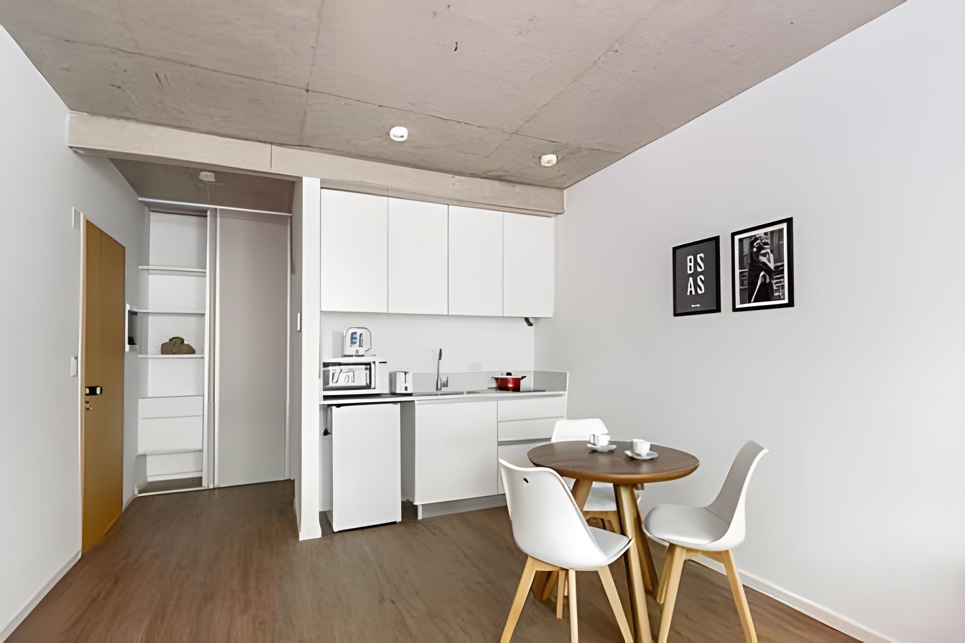 Une cuisine compacte avec des meubles blancs, un petit réfrigérateur et un coin repas comprenant deux chaises blanches et une table en bois.