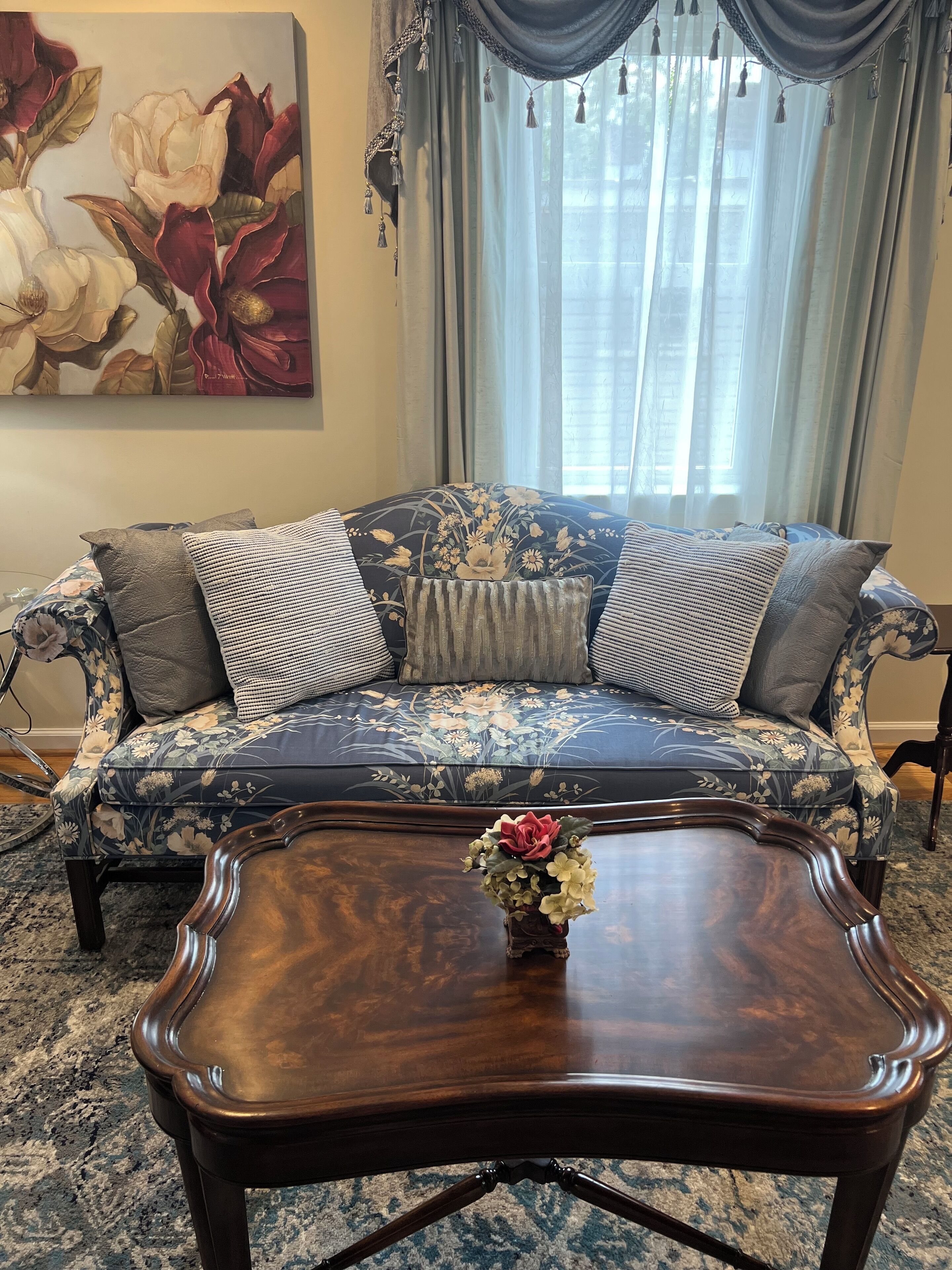 Una sala de estar con un sofá floral, una mesa de centro de madera con un jarrón de flores y un cuadro grande en la pared.