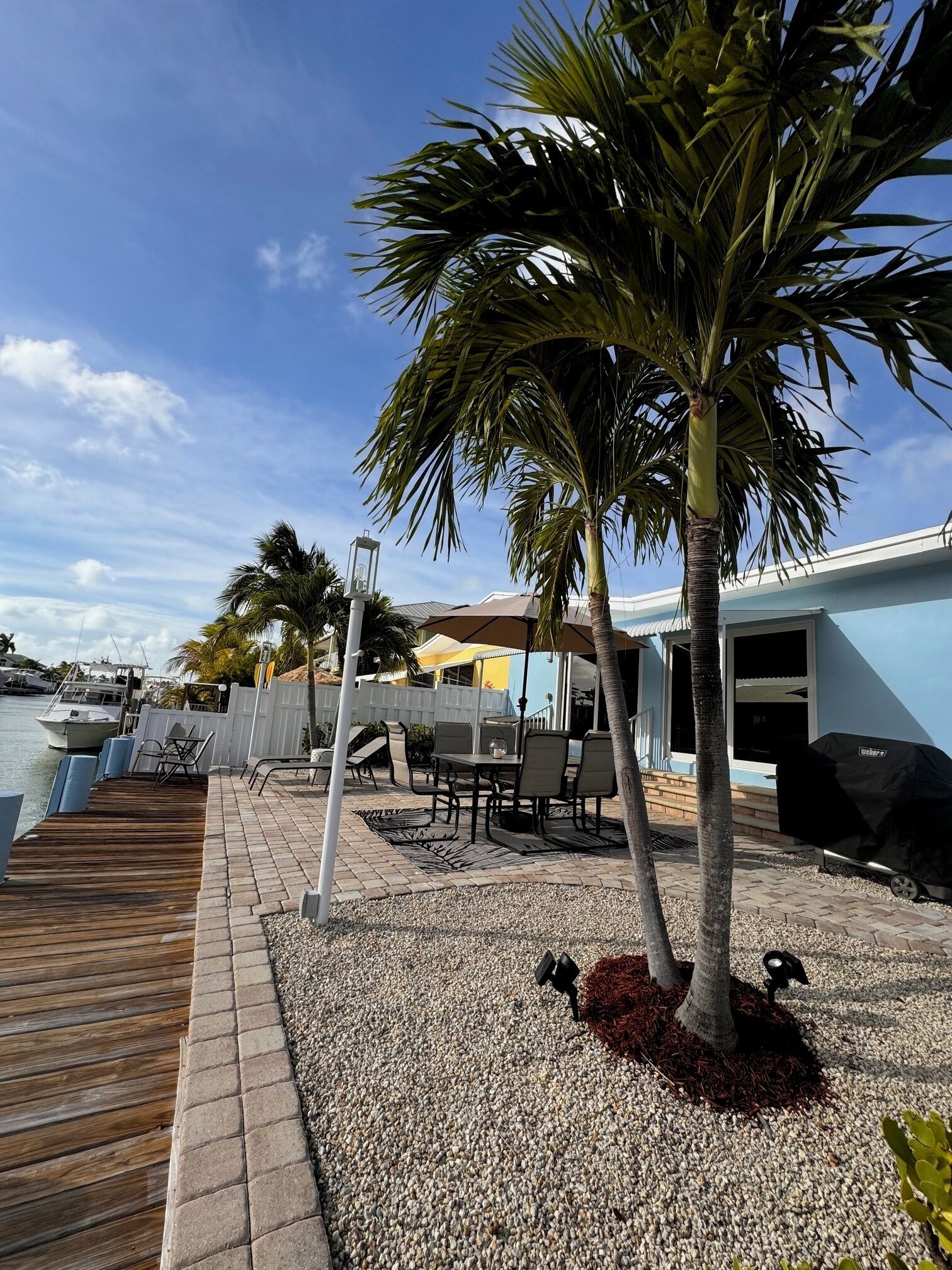 A patio area with a wooden deck, outdoor furniture, a palm tree, and a view of the marina with boats.