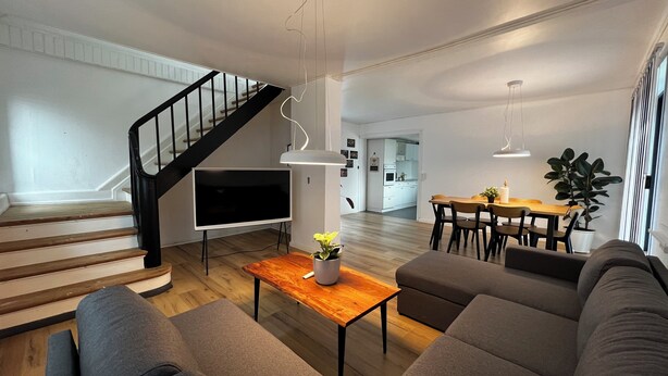 Ein modernes Wohnzimmer mit Treppe, einem Flachbildfernseher, einem Essbereich und einer Küche im Hintergrund.