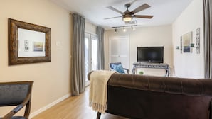 Una sala de estar con un sofá marrón, un televisor sobre un mueble negro, un ventilador de techo y un cuadro en la pared.
