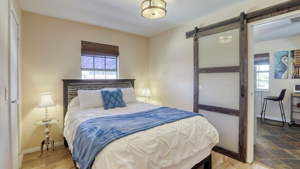Un dormitorio con cama, lámparas de noche, una ventana con persianas, una puerta con panel de vidrio y una lámpara en el techo.