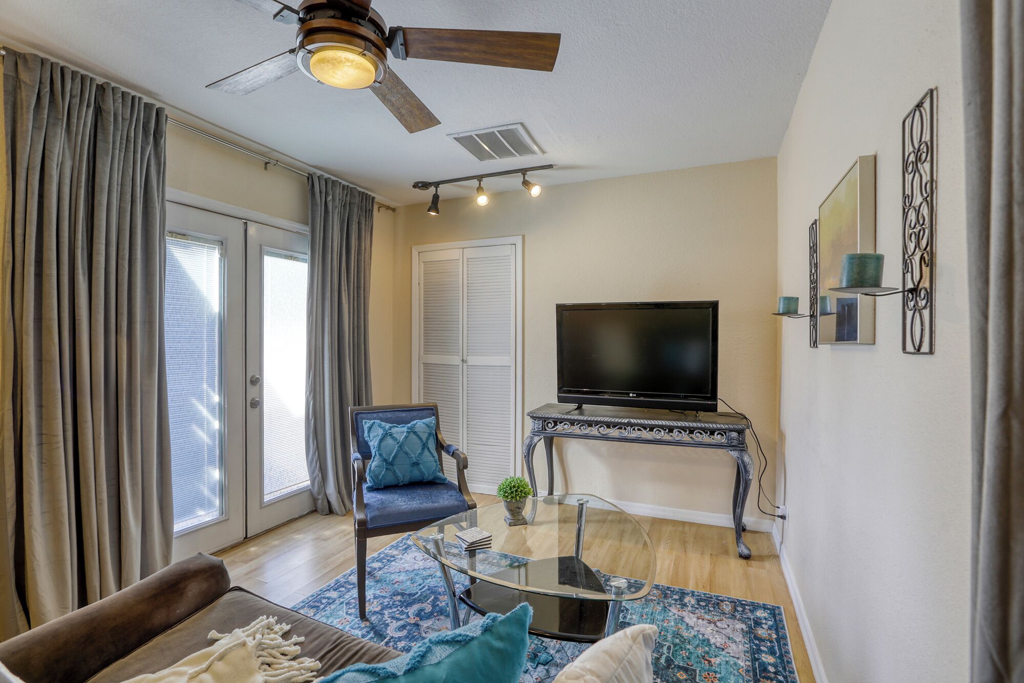 A living room with a ceiling fan, a flat-screen TV on a decorative stand, a glass coffee table, a sofa with cushions, and a patterned rug.