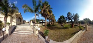 A courtyard with palm trees, a building with steps leading up to a door, and a well-maintained lawn. - Il Ciclamino (Melendugno)