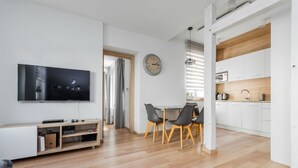 Una sala de estar moderna con una televisión de pantalla plana, piso de madera, un comedor con sillas y una cocina con armarios blancos.