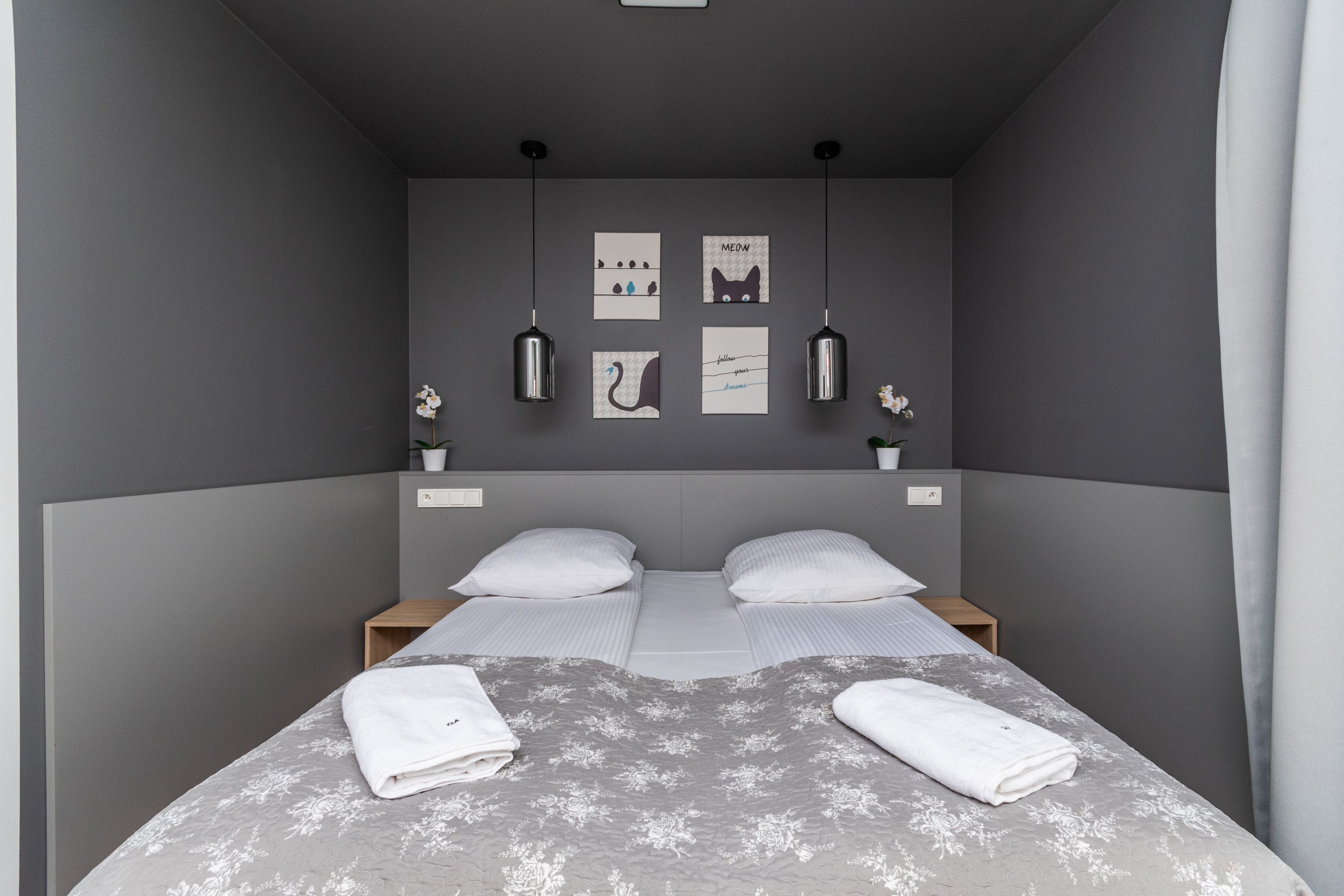 A modern hotel room with a grey wall, two beds with white linens, a grey floral bedspread, and artwork including a cat and a swan.