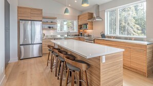 Une cuisine moderne dotée d’un grand îlot central, de tabourets de bar, d’appareils électroménagers en acier inoxydable et de grandes fenêtres offrant une vue sur l’extérieur.