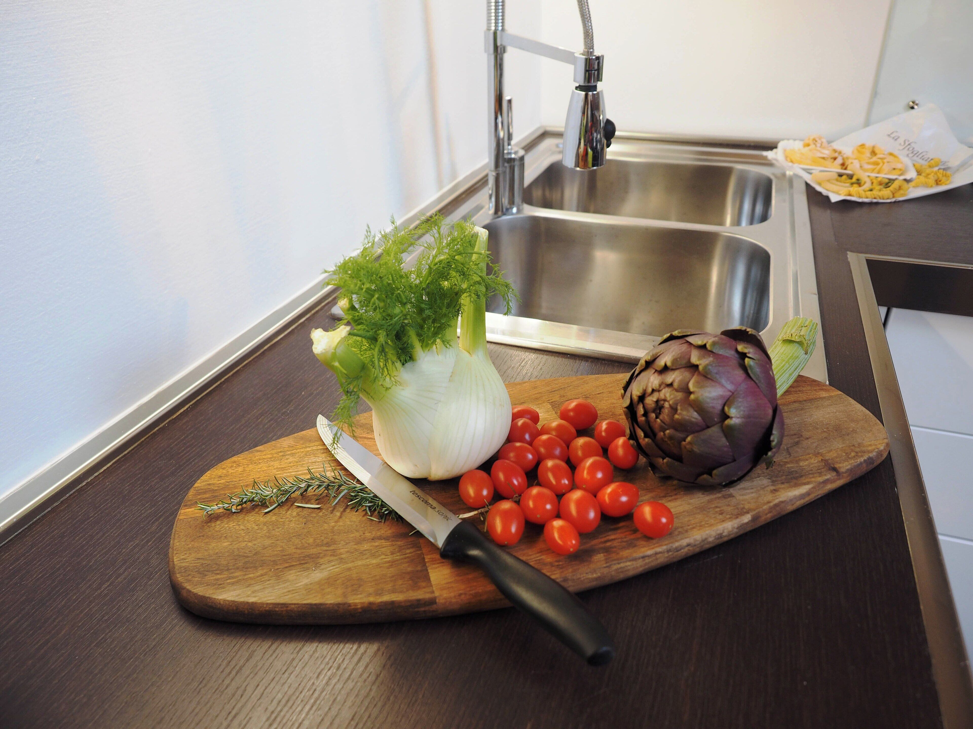 A kitchen counter with a wooden cutting board holding a knife, a head of cauliflower, cherry tomatoes, and an artichoke.