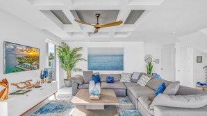 Ein modernes Wohnzimmer mit einer großen, grauen Eckcouch, einem Couchtisch und einem Deckenventilator. An den Wänden hängen zwei große Gemälde: eines zeigt einen Sonnenuntergang über dem Wasser, das andere einen blauen Himmel.