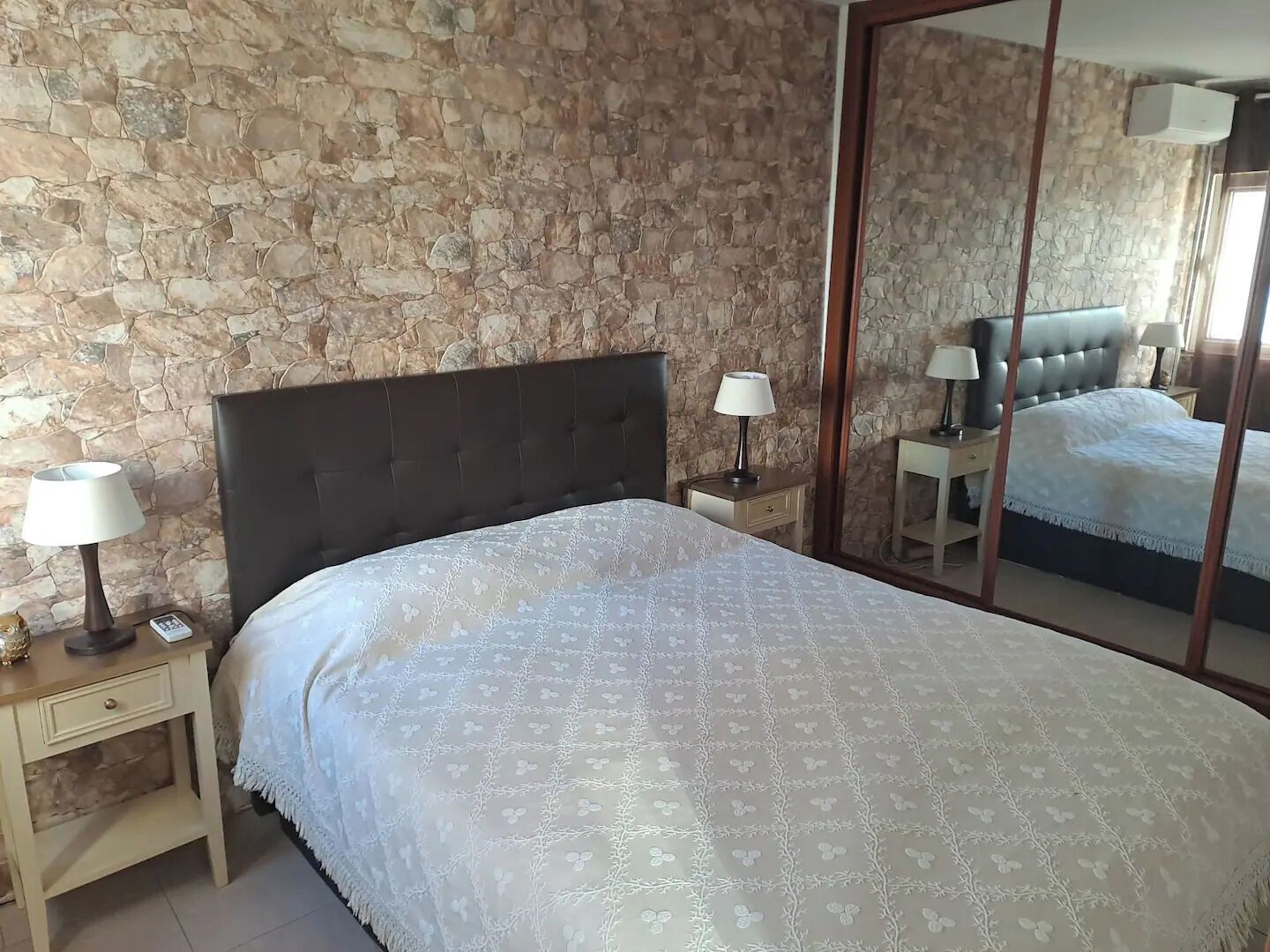 A bedroom with a stone wall, a bed with a patterned bedspread, two bedside tables with lamps, and a large mirror.