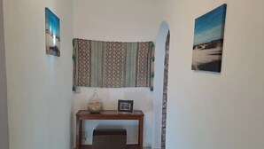 A hallway with a patterned tapestry hanging on the wall, a small wooden console table with a glass jar and a framed picture, and a tiled floor.