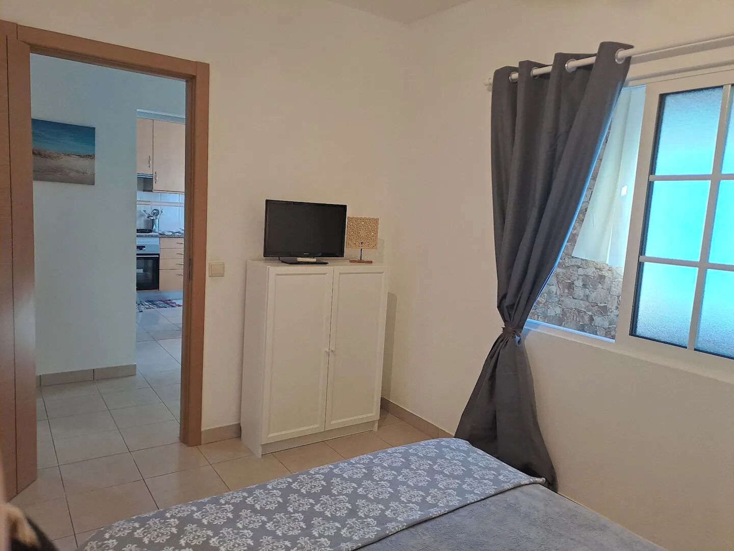 A bedroom with a bed, a TV on a cabinet, a window with curtains, and a view of a kitchen and dining area.