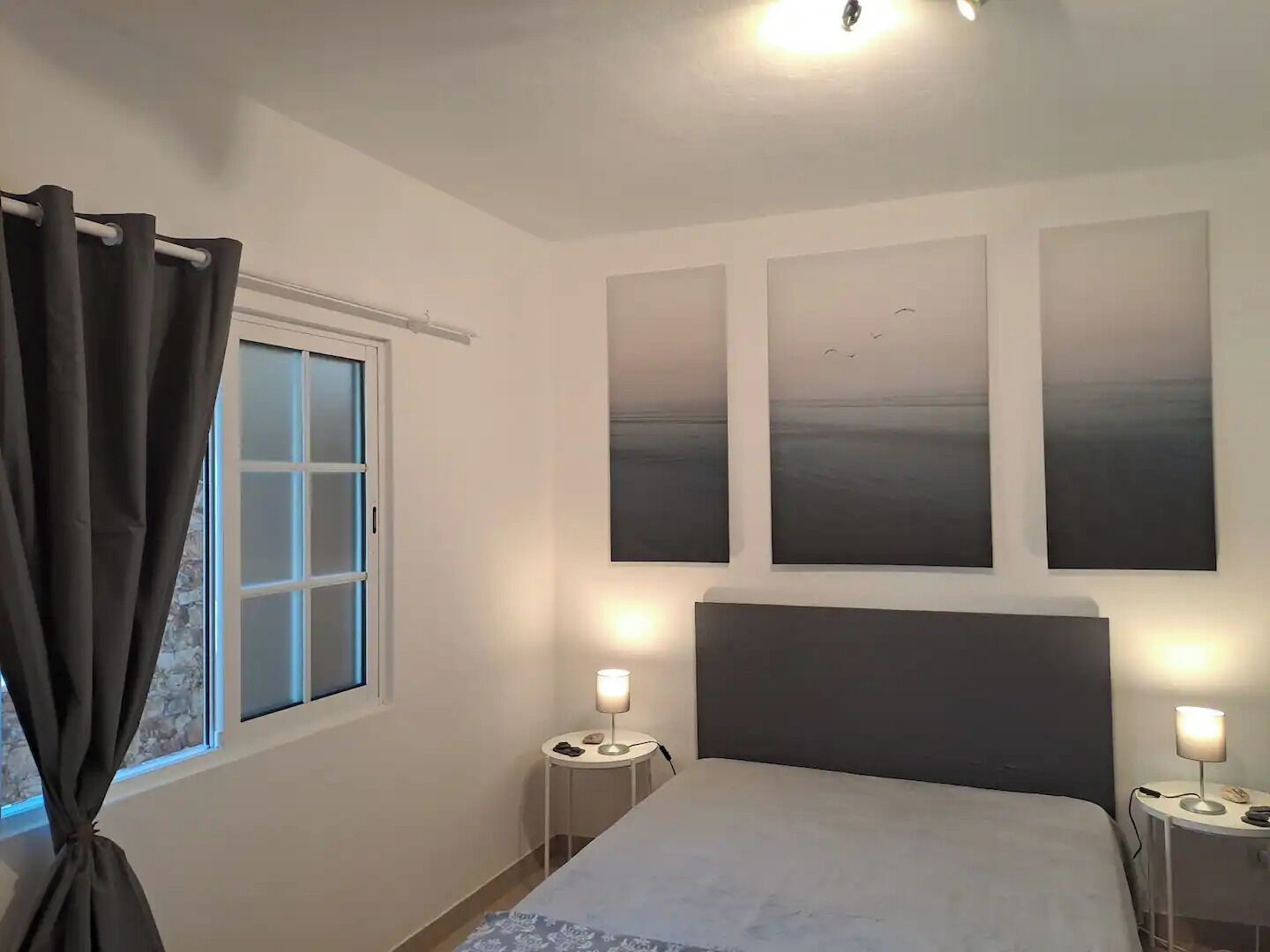 A bedroom with a bed, two bedside tables with lamps, a window with curtains, and three wall-mounted paintings.