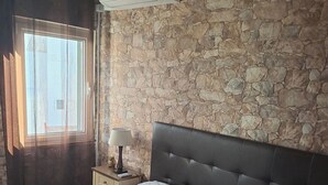 A bedroom with a stone wall, a bed with a quilt, a nightstand with a lamp, and an air conditioning unit.