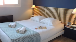 Una cama bien hecha con sábanas blancas y un edredón estampado, una mesita de noche con una lámpara, y una ventana con persianas.