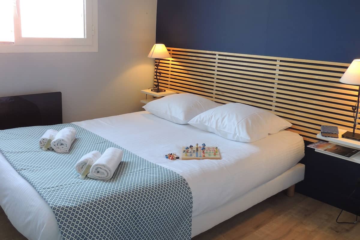 Una cama bien hecha con sábanas blancas y un edredón estampado, una mesita de noche con una lámpara, y una ventana con persianas.