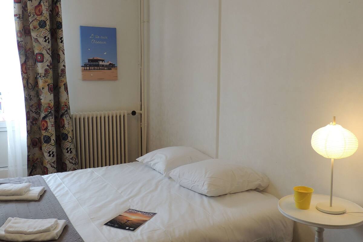 Una cama bien hecha con almohadas, una mesita de noche con lámpara, un radiador y un cartel en la pared.