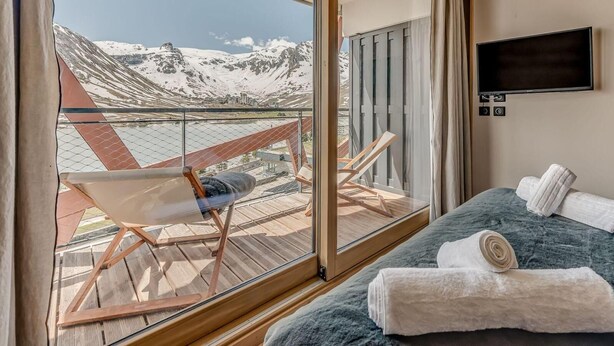 Habitación de hotel con un ventanal grande que ofrece vistas a la montaña, una cama con dos toallas, un televisor en la pared y una silla junto a la ventana.