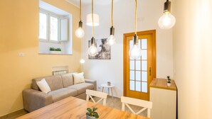 A cozy living room with a wooden dining table, a sofa, and a small side table. There are hanging light fixtures and a window with a view. - Smooth Vibes at Chiaia by Napoliapartments (Naples)