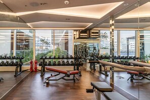 Un gimnasio con diversos equipos de ejercicio, ventanas amplias y vista a edificios exteriores.