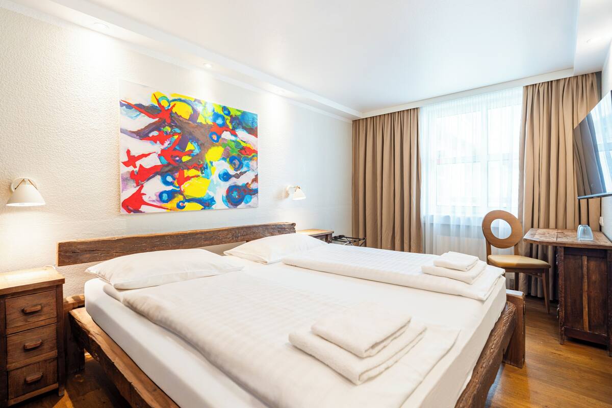 Habitación de hotel con dos camas, un escritorio, una silla, un televisor y un cuadro abstracto de colores en la pared.