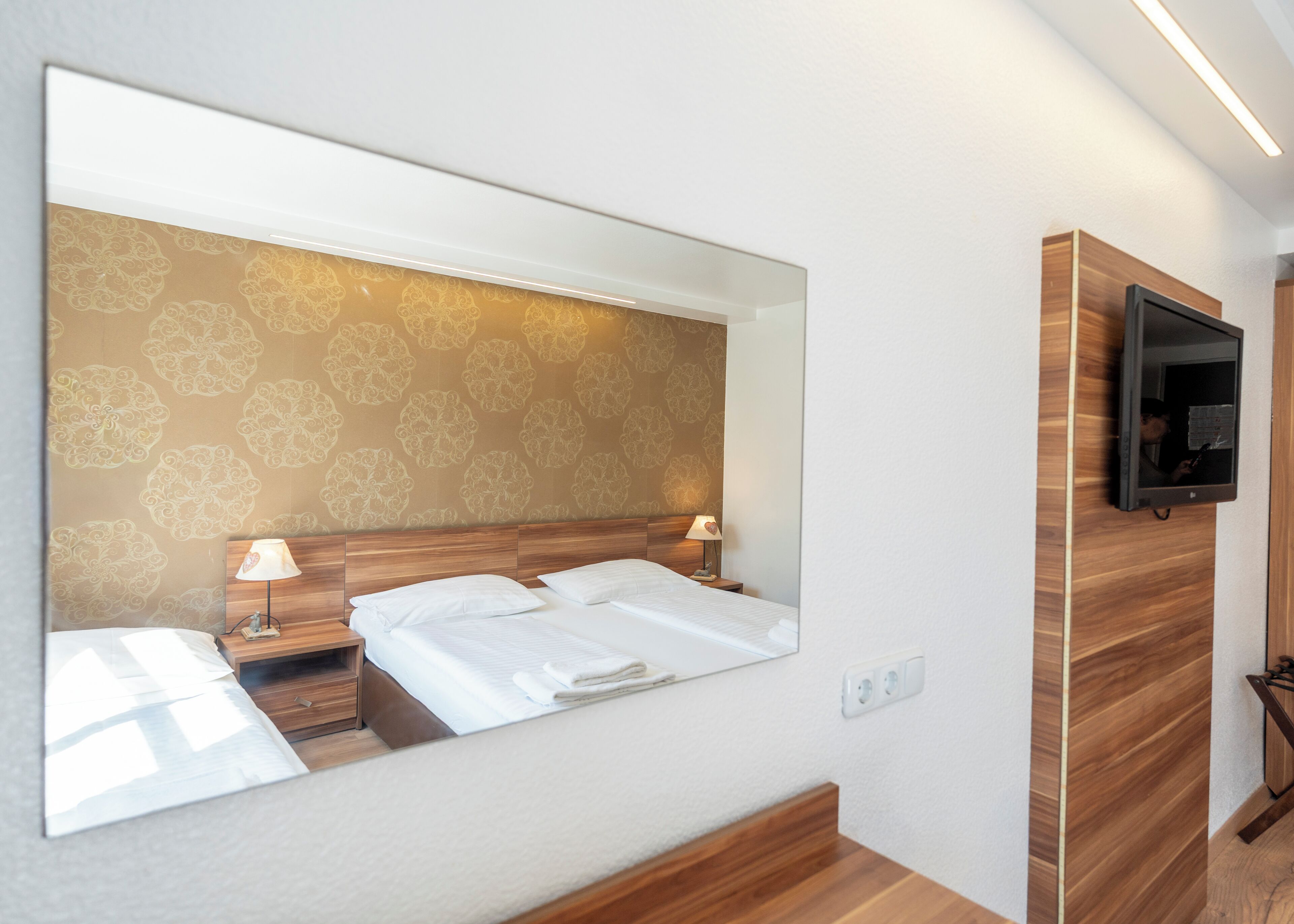 Ein Hotelzimmer mit einem großen Spiegel, einem Bett mit weißer Bettwäsche, einem Holzkopfende, einem Nachttisch mit Lampe und einem an der Wand befestigten Fernseher.