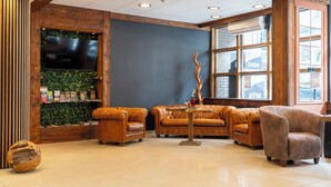 Een lobby met een hoge plafond, houten balken, een receptiebalie, leren banken, een bijzettafeltje en een groot raam met een geldautomaat.