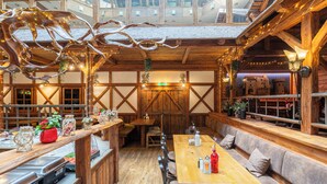 A rustic interior with wooden beams, a high ceiling, and a central dining area with benches and tables.
