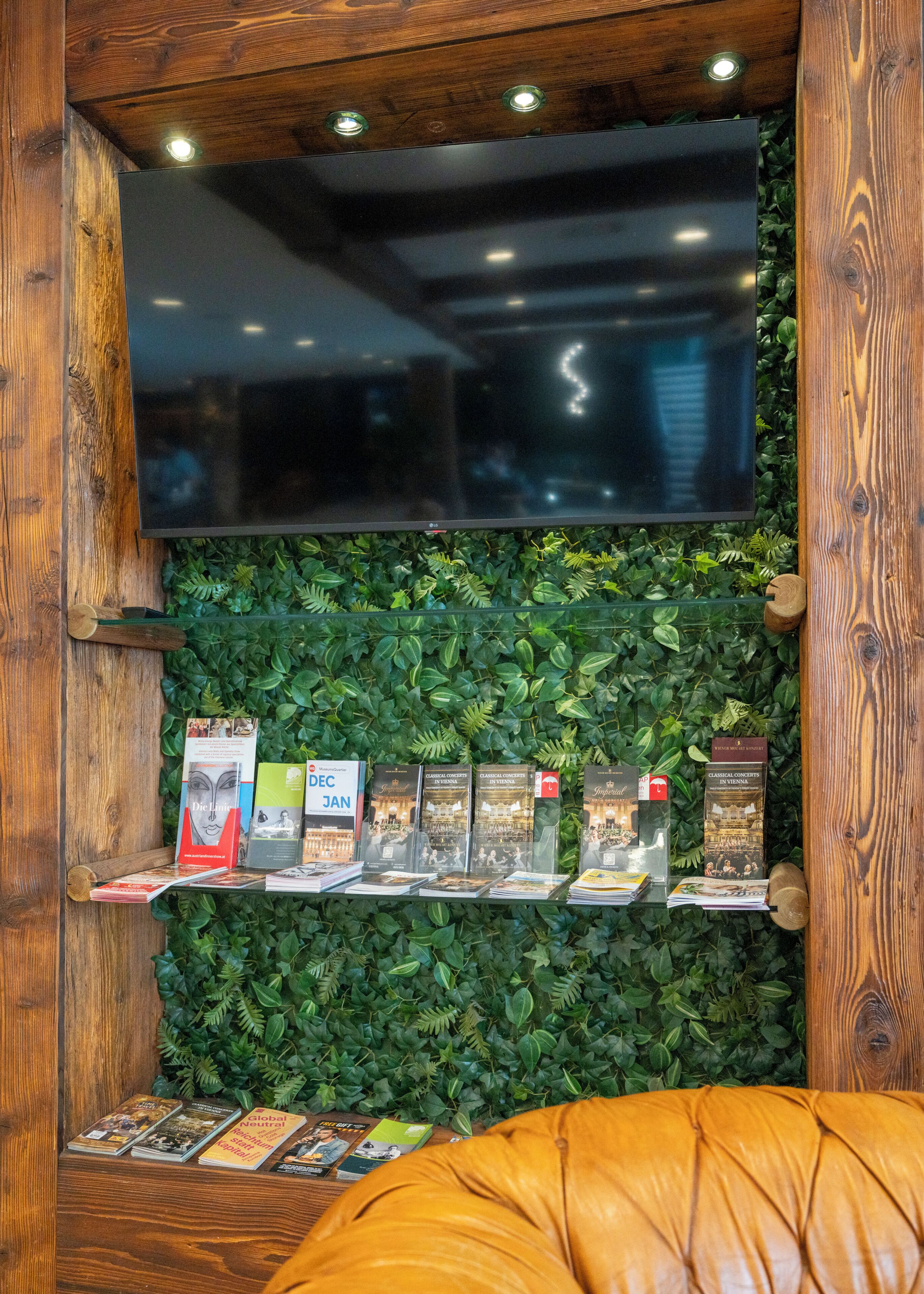 A room with a large flat-screen TV mounted on a wooden wall, surrounded by a green leafy wall. There are books and magazines on a shelf below the TV.