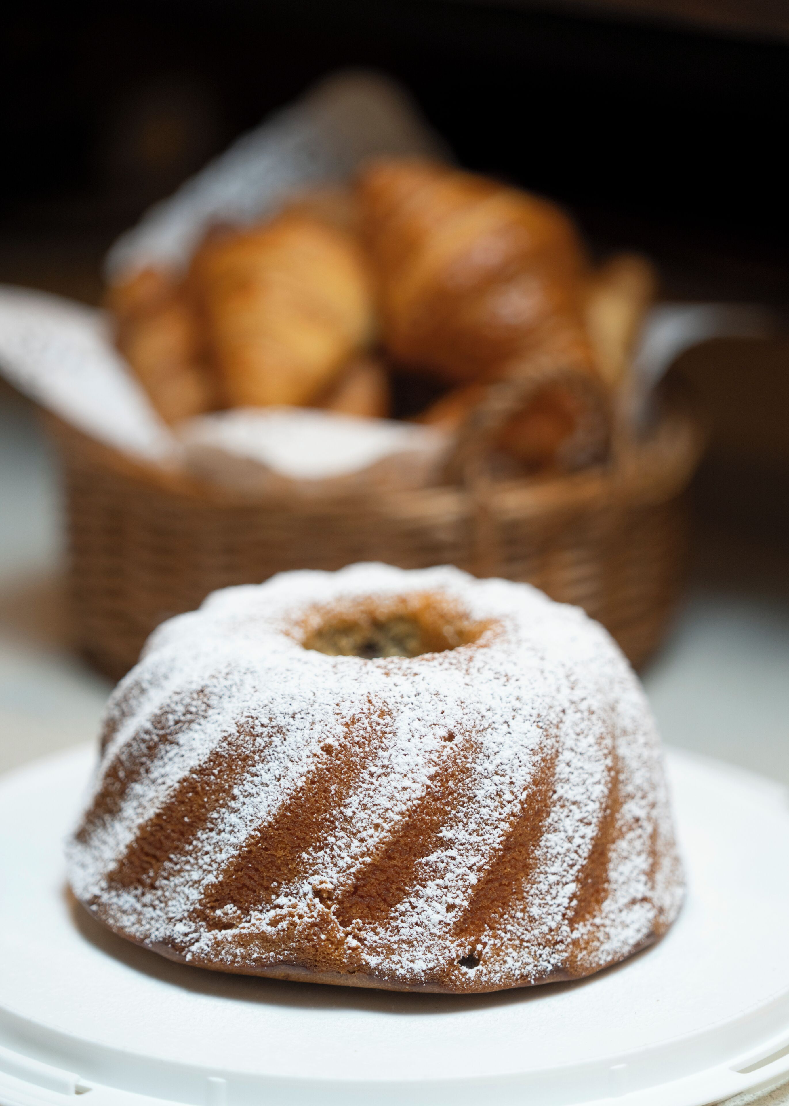 A bundt cake on a plate with powdered sugar, and a basket of croissants in the background.