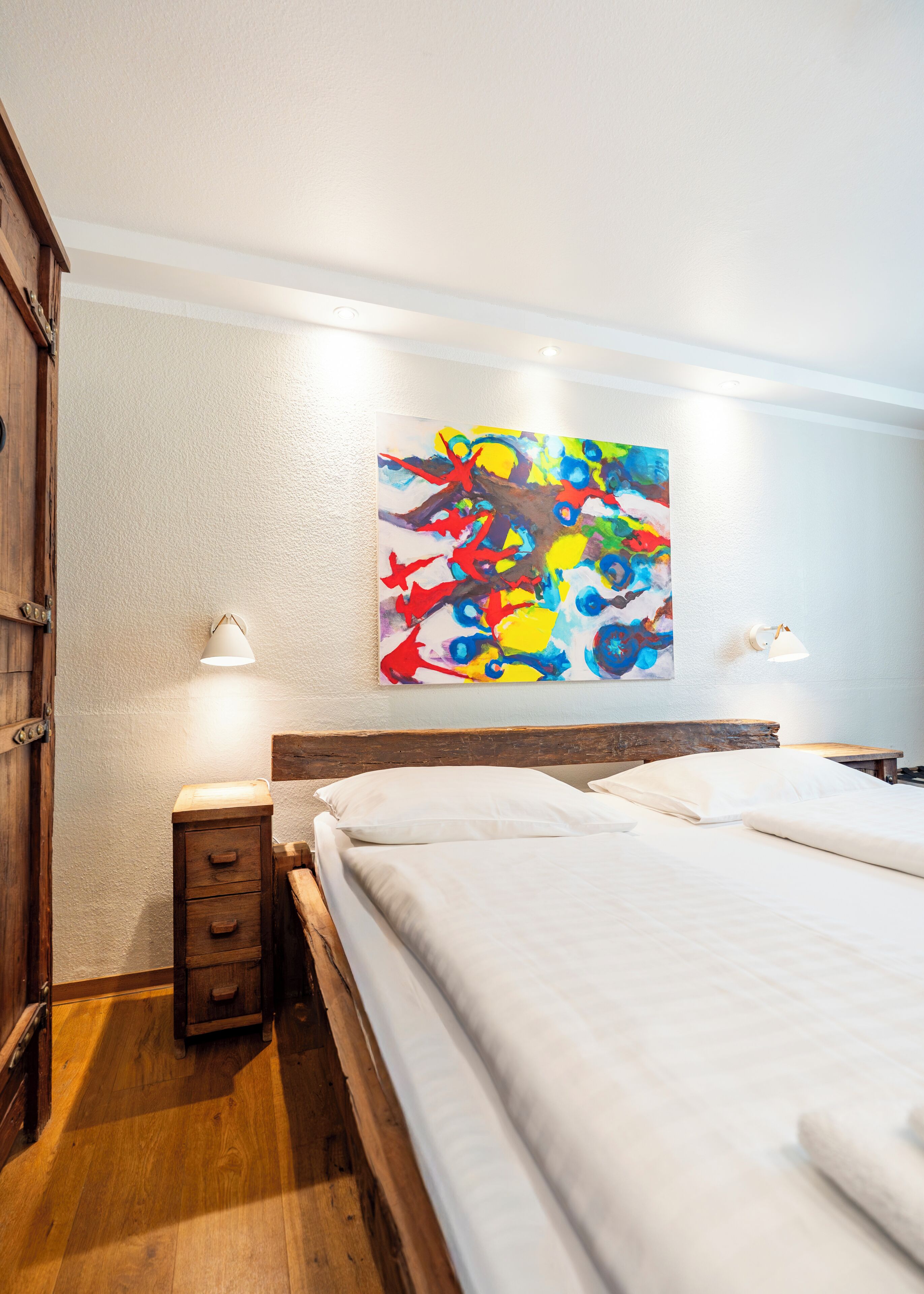 Habitación de hotel con una cama de madera, una mesita de noche, una lámpara y un cuadro abstracto de colores en la pared.