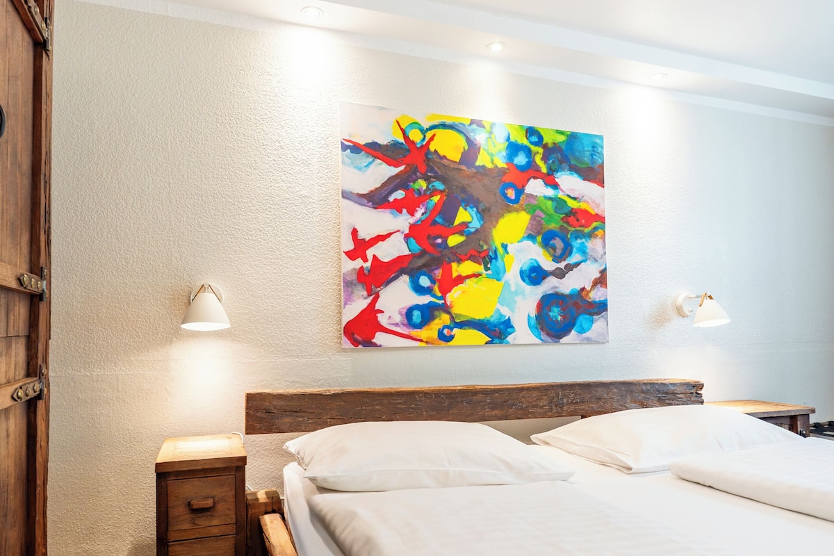 Habitación de hotel con una cama de madera, una mesita de noche, una lámpara y un cuadro abstracto de colores en la pared.