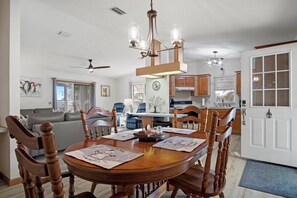 A dining area with a wooden table, chairs, and a chandelier. There is a kitchen in the background with wooden cabinets and a window.