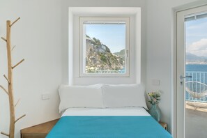 Une chambre avec un lit, une fenêtre donnant sur la côte, un porte-manteau en bois et une chaise à l’extérieur.