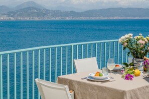 Une table dressée avec des assiettes, des verres et des fleurs, sur un balcon donnant sur une étendue d’eau et des montagnes lointaines.