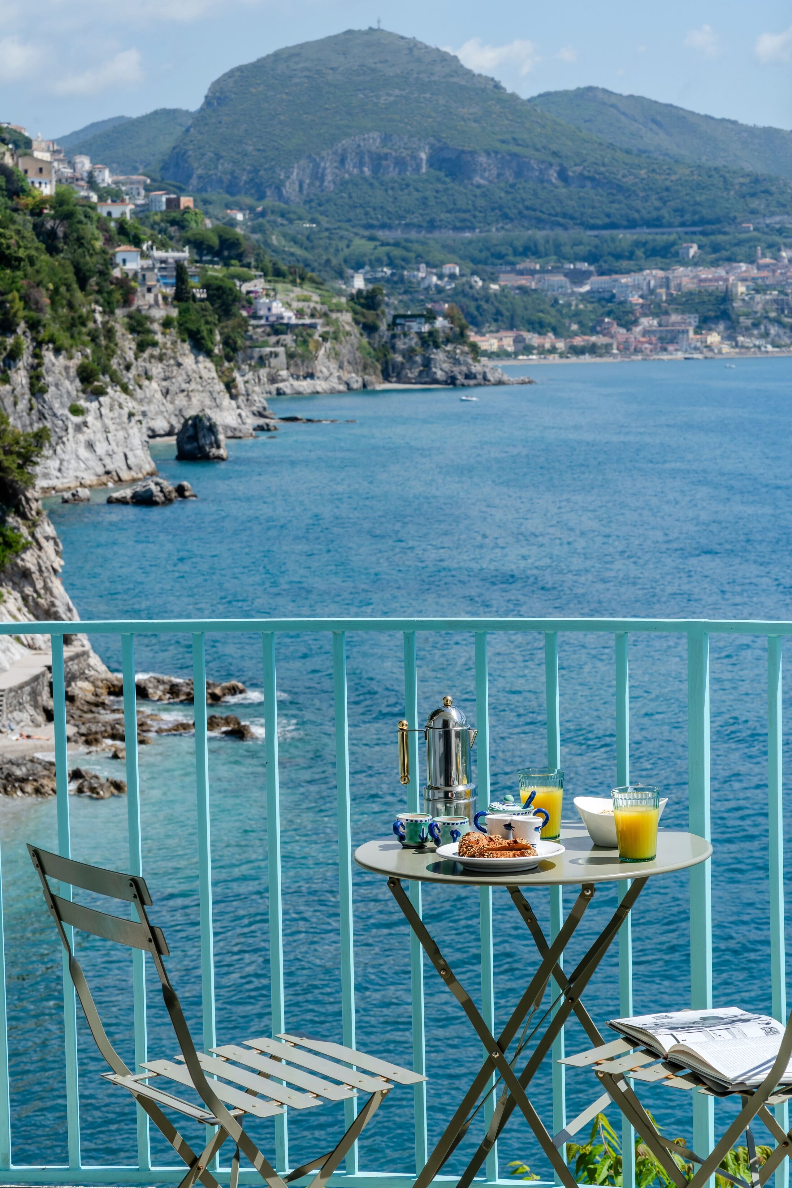 A balcony with a table set for breakfast overlooking a coastal town and the sea.