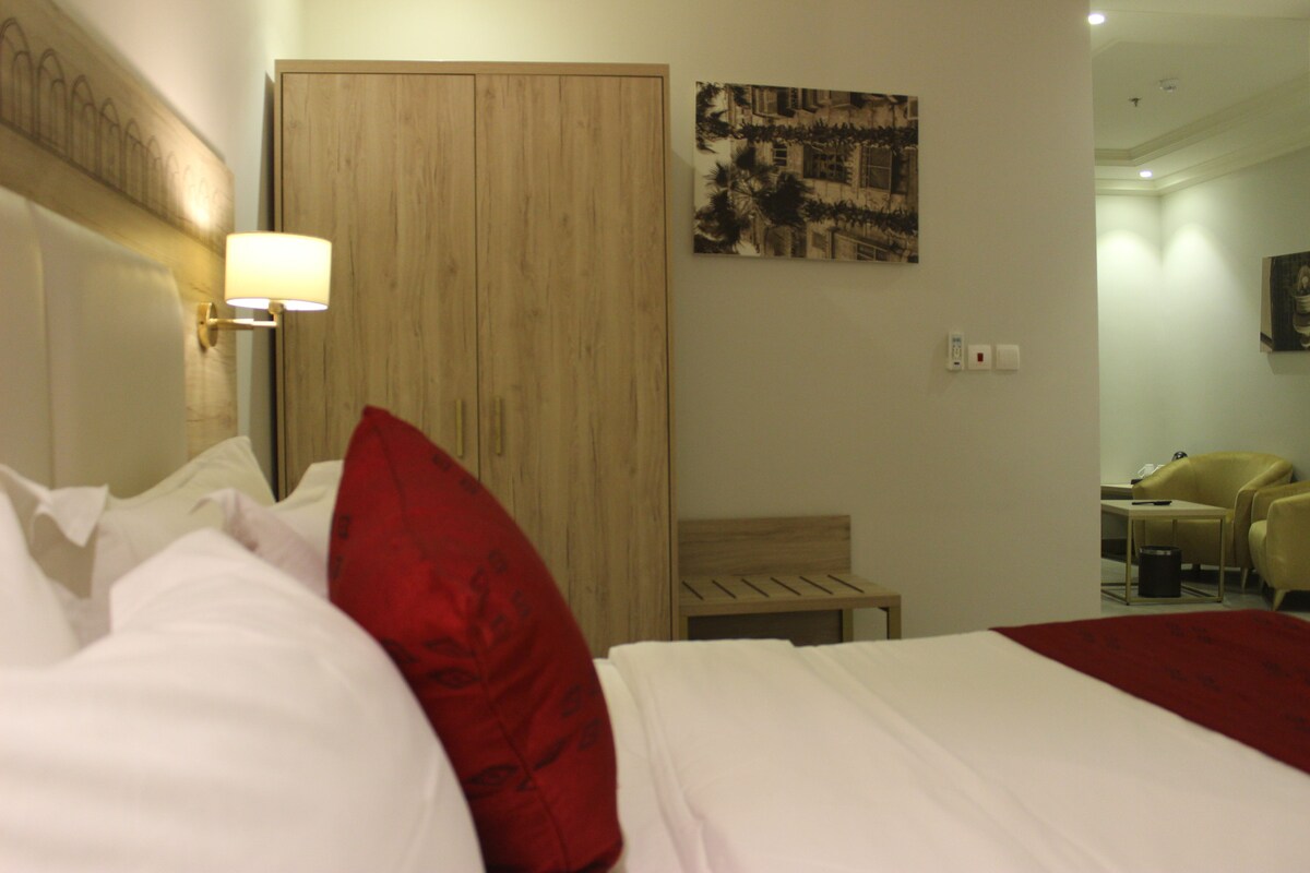 Een hotelkamer met een bed, een houten garderobe, een wandlamp, een klein tafeltje en een stoel.