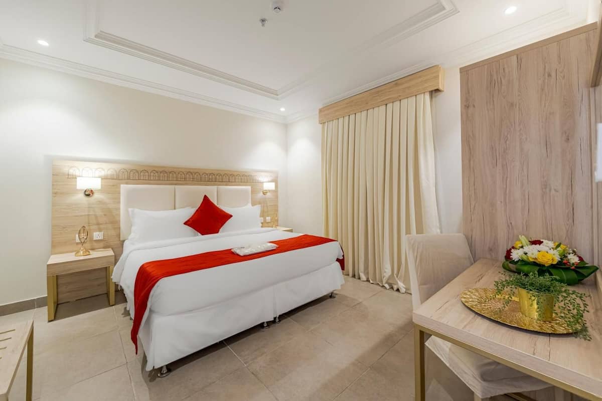 Habitación de hotel con una cama grande, una mesita de noche con lámpara, cabecero de madera, ventana con cortinas y una pequeña mesa con un jarrón de flores.