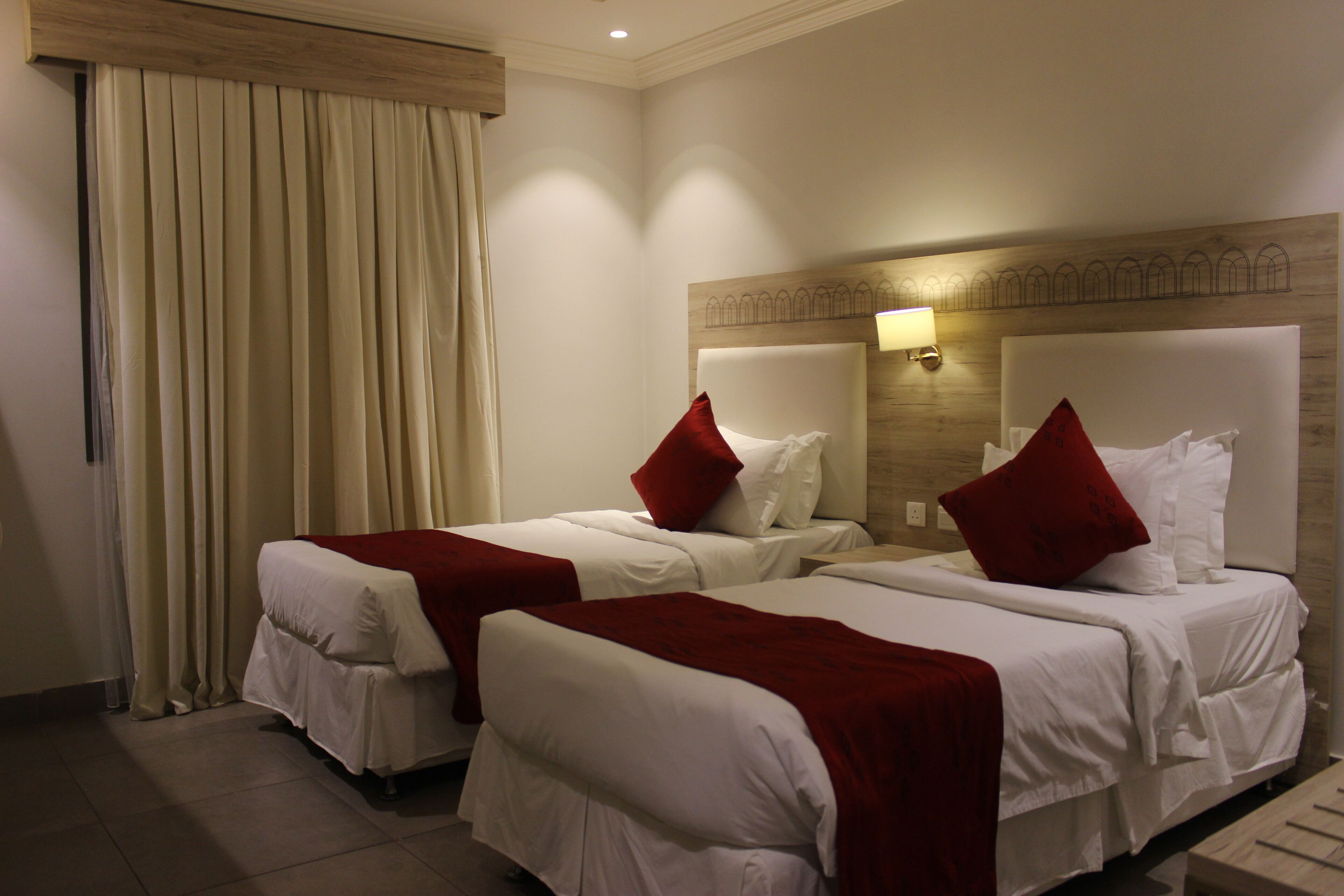 A hotel room with two beds, each with white bedding and red pillows. There is a wooden headboard with a light fixture above the beds.
